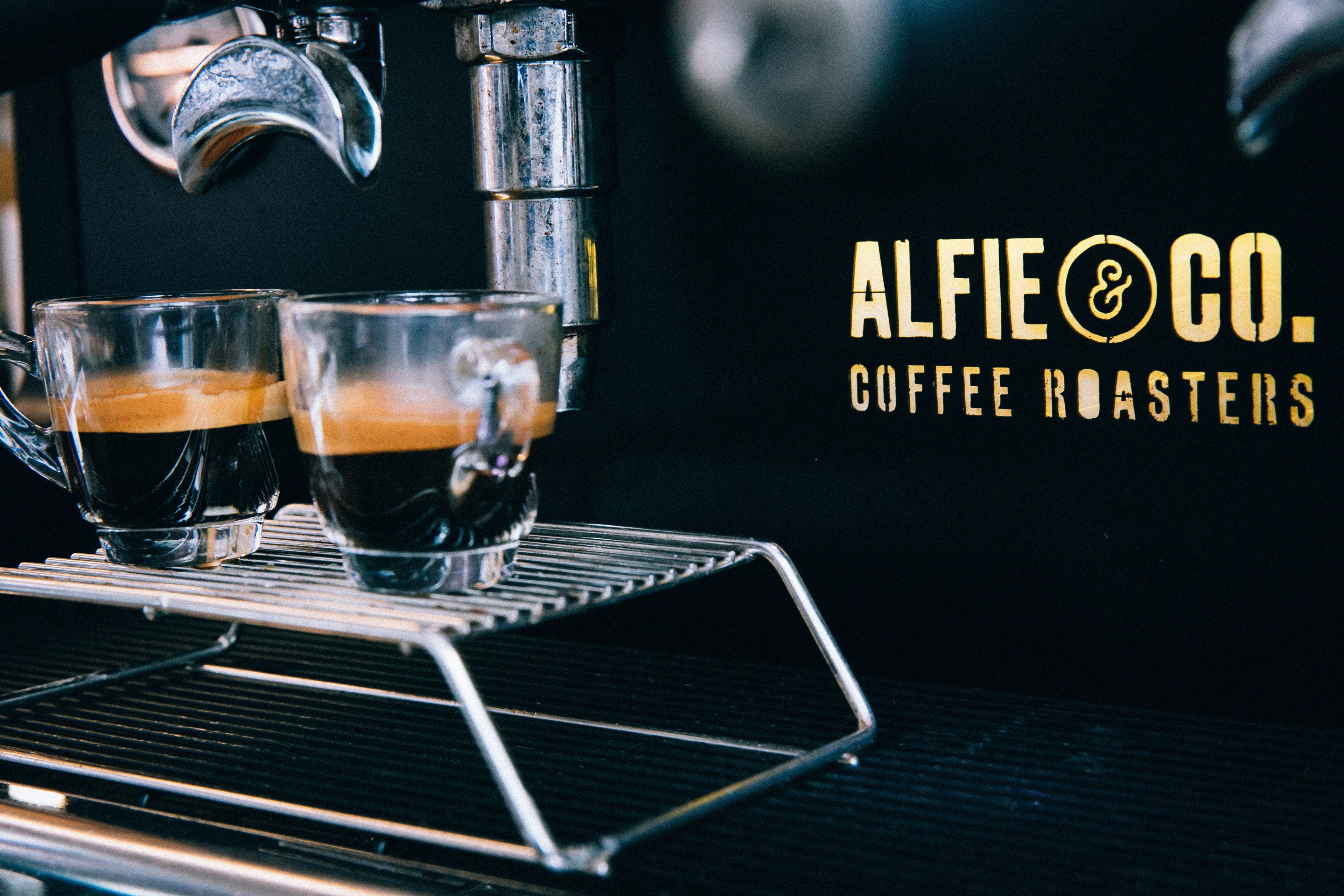 Two clear glass cups filled with freshly brewed espresso sit on a chrome drip tray of a professional espresso machine. The "ALFIE & CO. COFFEE ROASTERS" logo is visible in gold lettering on a black background. Image highlights small batch, hand-roasted coffee from Alfie & Co, based in Killearn, Scotland, available for UK-wide online purchase at www.alfiecoffee.co.uk.