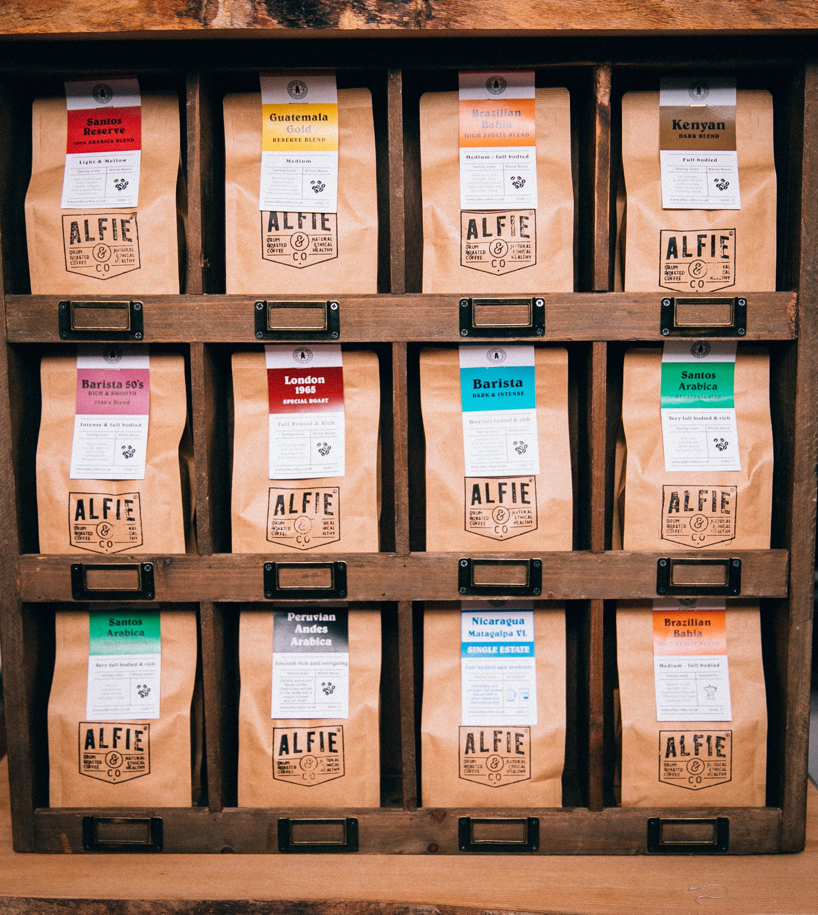 Twelve kraft paper bags of Alfie & Co. coffee beans displayed in a vintage wooden drawer unit with label slots. Each bag is labeled with a different origin or blend name, including "Guatemala," "Kenyan," "London 1965," and "Brazilian Bahia."  The Alfie & Co. logo is prominently featured on each bag, showcasing our wide selection of hand-roasted, small-batch coffees available for UK-wide delivery from Killearn, Scotland at www.alfiecoffee.co.uk.  Highlights the variety and unique character of our coffee offe