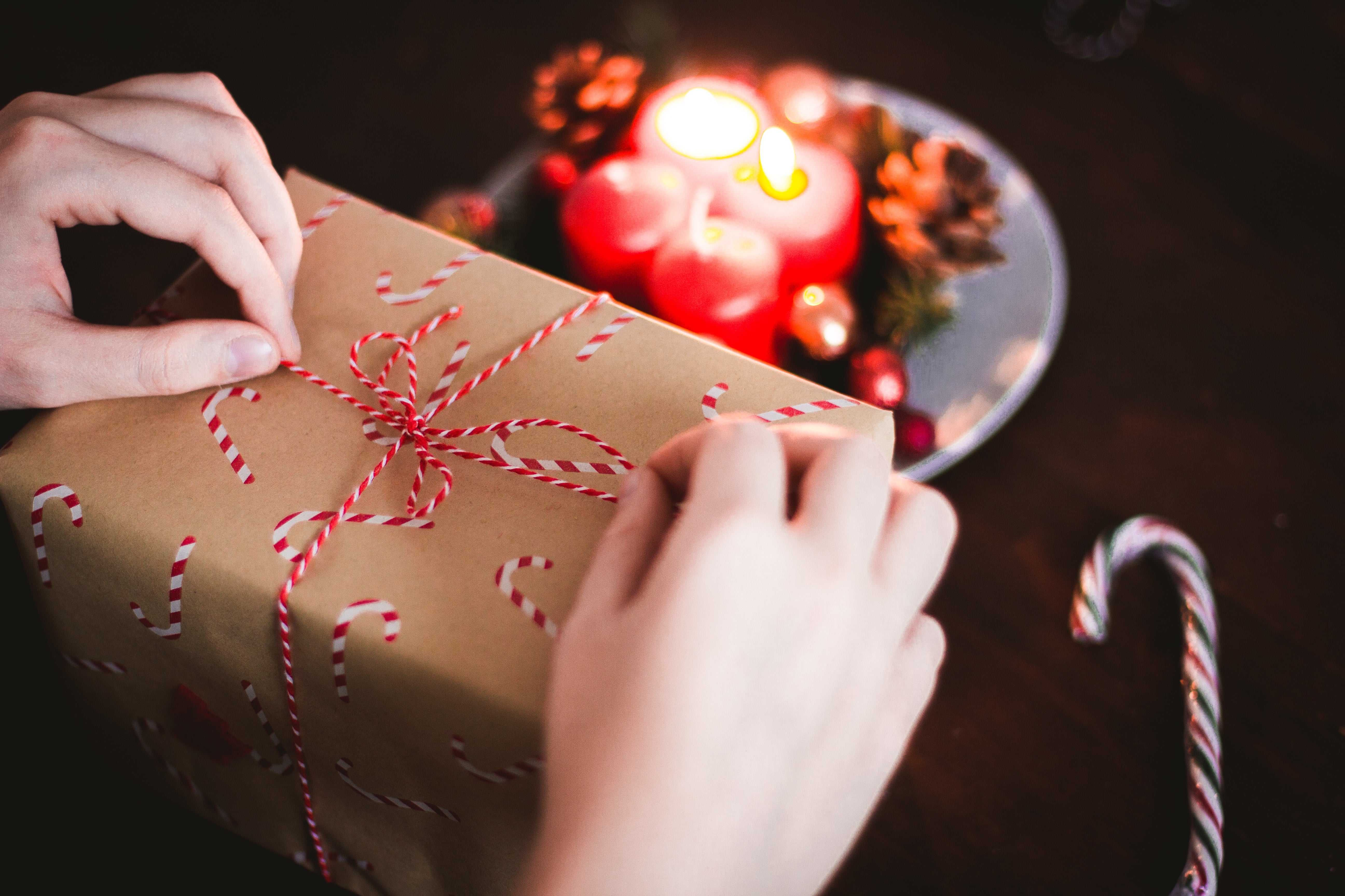 Hands tying a red and white string around a brown paper-wrapped gift box with a candy cane pattern. A blurred plate with lit red candles and pine cones is visible in the background, creating a warm, festive atmosphere.  Perfect for gifting Alfie & Co. coffee during the holiday season.  Share the warmth of our hand-roasted, small-batch coffee from Killearn, Scotland, available for UK delivery at www.alfiecoffee.co.uk.  Highlights the cozy and thoughtful nature of our coffee gifts.
