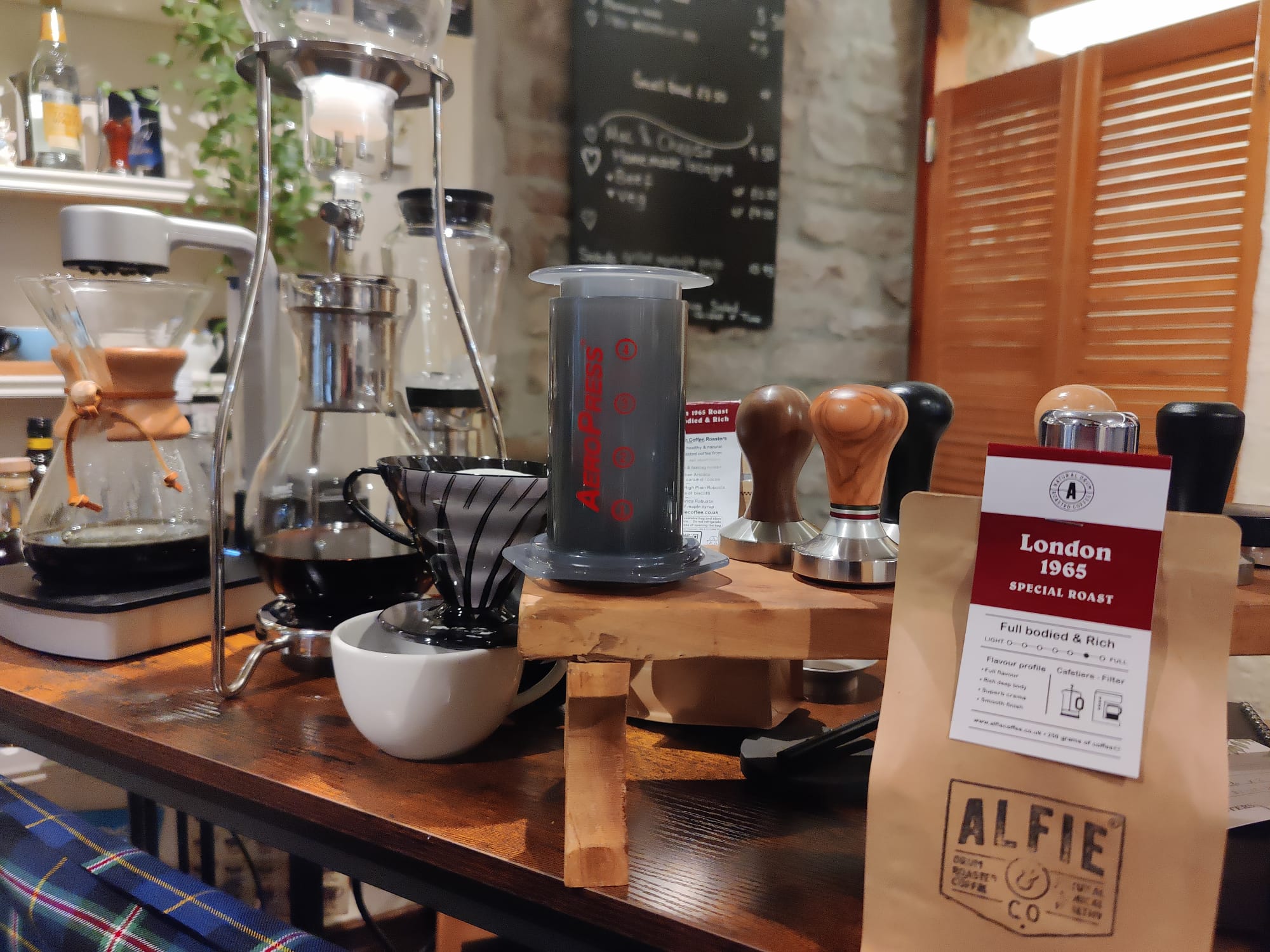 Alfie & Co. London 1965 Special Roast coffee bag displayed alongside various brewing equipment, including a Chemex, Aeropress, and pour-over setup, on a wooden counter.  This scene showcases a hands-on coffee experience session, highlighting the versatility and quality of our hand-roasted beans.  Learn to brew like a barista and discover the rich, full-bodied flavor of our coffee, available for UK delivery from Killearn, Scotland at www.alfiecoffee.co.uk.  Illustrates the educational and interactive aspect 