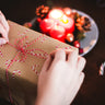  Hands tying a red and white string around a brown paper-wrapped gift box with a candy cane pattern. A blurred plate with lit red candles and pine cones is visible in the background, creating a warm, festive atmosphere.  Perfect for gifting Alfie & Co. coffee during the holiday season.  Share the warmth of our hand-roasted, small-batch coffee from Killearn, Scotland, available for UK delivery at www.alfiecoffee.co.uk.  Highlights the cozy and thoughtful nature of our coffee gifts.